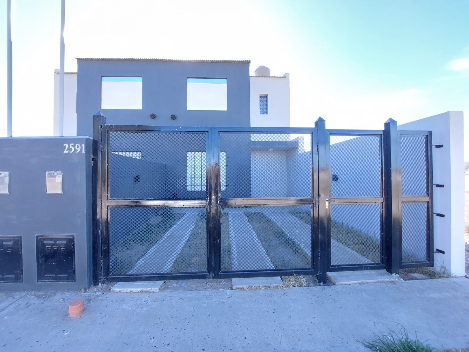 La imagen muestra una fachada moderna de una propiedad residencial de dos pisos. El ambiente es exterior, con un diseño contemporáneo. La estructura parece estar en buen estado de conservación. Los materiales visibles incluyen concreto, metal y vidrio. La puerta y el portón son de metal negro con detalles de malla, brindando seguridad y un estilo moderno. La iluminación natural es abundante, gracias a las amplias ventanas en el segundo piso. Destaca el camino de entrada con césped a los lados, que conduce al interior de la propiedad. La fachada tiene un acabado en tonos grises, lo que le da un aspecto elegante y sobrio.