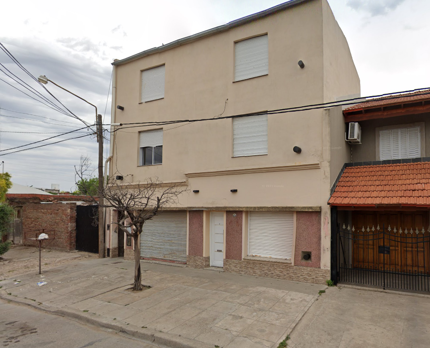 La imagen muestra un edificio residencial de tres pisos en una zona urbana. El exterior está en buen estado de conservación, con paredes de color beige y detalles en piedra en la planta baja. Las ventanas tienen persianas enrollables blancas, lo que sugiere un buen control de la iluminación natural. La entrada principal es sencilla, con una puerta blanca. No se observan jardines ni áreas verdes, y la acera es de cemento. La iluminación exterior parece adecuada, con varias luces instaladas en la fachada. Destaca la presencia de un árbol pequeño frente al edificio, aunque sin follaje visible.