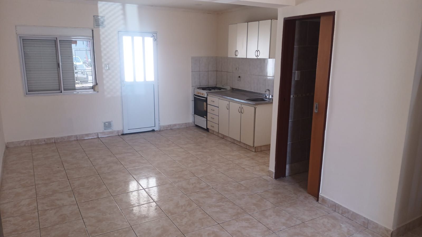 La imagen muestra un ambiente tipo cocina-comedor integrado. El estado de conservación parece bueno, con paredes y techo en buen estado. El piso es de cerámica en tonos beige, lo que facilita la limpieza. La cocina cuenta con muebles blancos, una encimera de acero inoxidable y un horno a gas. Las paredes de la cocina están revestidas con azulejos claros. La iluminación natural proviene de una ventana y una puerta con vidrio, ambas con persianas. Destaca la amplitud del espacio y su luminosidad. La puerta de madera a la derecha parece conducir a otra habitación o baño.